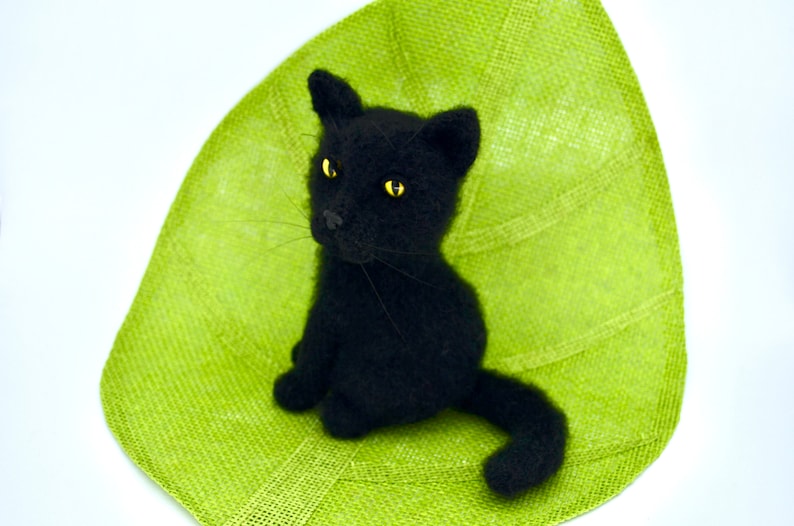 May include: A black crocheted cat with yellow eyes sitting on a green textured surface.