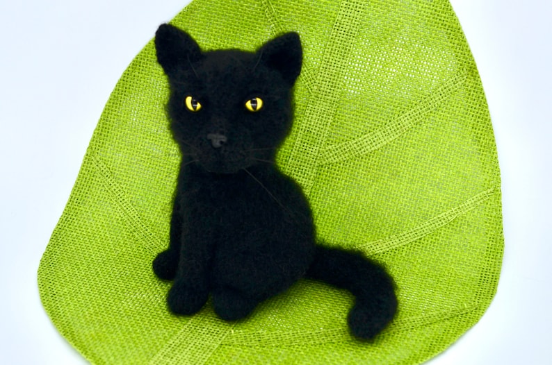 May include: A black knitted cat figurine with yellow eyes sitting on a green burlap surface.