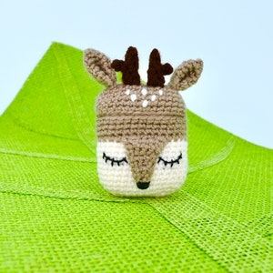 May include: A handmade crochet deer toy with closed eyes, brown antlers, and a white and brown face. The deer is set against a bright green textured background. The toy is likely a decorative item or a child's toy.