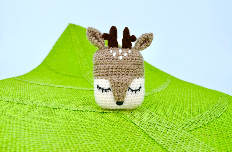 May include: A handmade crochet deer toy with closed eyes, brown antlers, and a white and brown face. The deer is set against a bright green textured background. The toy is likely a decorative item or a child's toy.