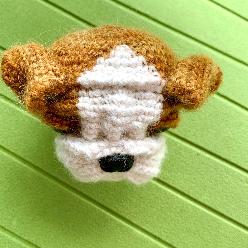 May include: A crocheted bulldog head with brown and white fur. The bulldog has a black nose and white fur around its eyes.