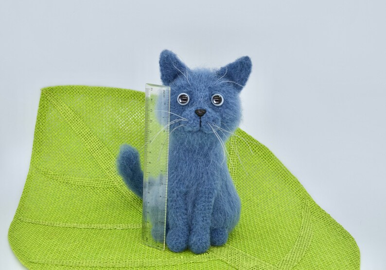 May include: A blue knitted cat toy with white whiskers and large black eyes. The cat is sitting on a green woven mat.
