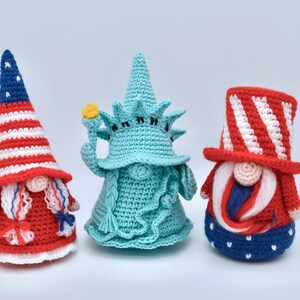 Puede incluir: Tres gnomos de crochet, cada uno representando un icono americano diferente. El primer gnomo lleva un sombrero a rayas rojo, blanco y azul y un vestido rojo y blanco. El segundo gnomo tiene la forma de la Estatua de la Libertad, con un cuerpo de crochet verde y una antorcha amarilla. El tercer gnomo lleva un sombrero de copa a rayas rojo, blanco y azul y un vestido a rayas azul y blanco.