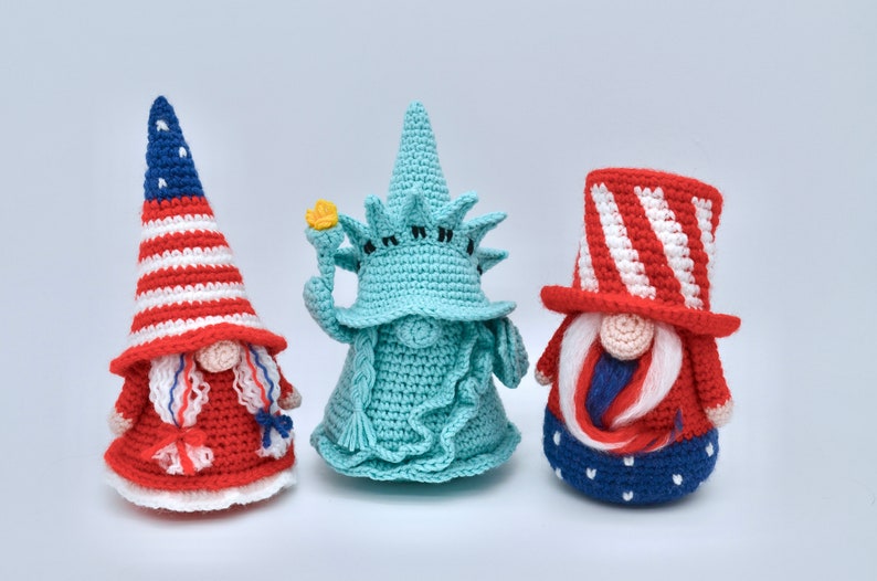 Puede incluir: Tres gnomos de crochet, cada uno representando un icono americano diferente. El primer gnomo lleva un sombrero a rayas rojo, blanco y azul y un vestido rojo y blanco. El segundo gnomo tiene la forma de la Estatua de la Libertad, con un cuerpo de crochet verde y una antorcha amarilla. El tercer gnomo lleva un sombrero de copa a rayas rojo, blanco y azul y un vestido a rayas azul y blanco.