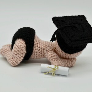 May include: A crocheted baby doll wearing a black graduation cap and a diaper. The doll is holding a rolled up diploma with a gold ribbon.