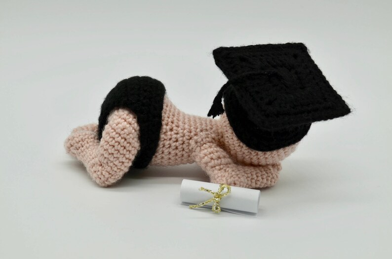 May include: A crocheted baby doll wearing a black graduation cap and a diaper. The doll is holding a rolled up diploma with a gold ribbon.