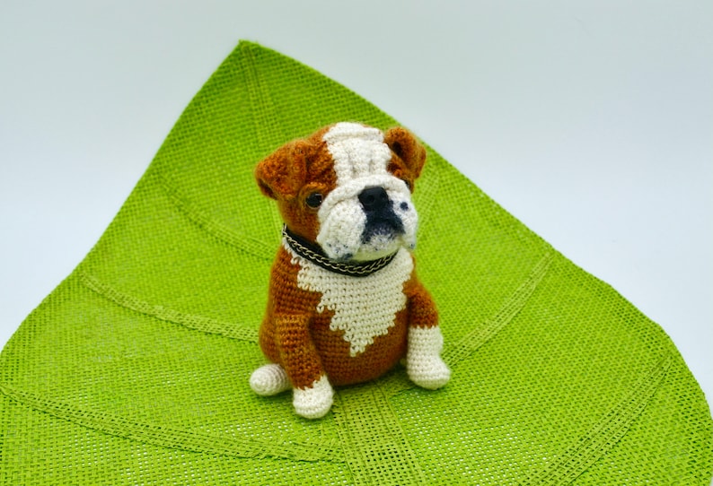 May include: A brown and white crocheted bulldog sitting on a green burlap surface.