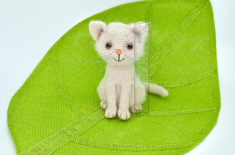 May include: A white crocheted cat with brown eyes and a pink nose sits on a green leaf-shaped mat. The cat is about 6 inches tall.