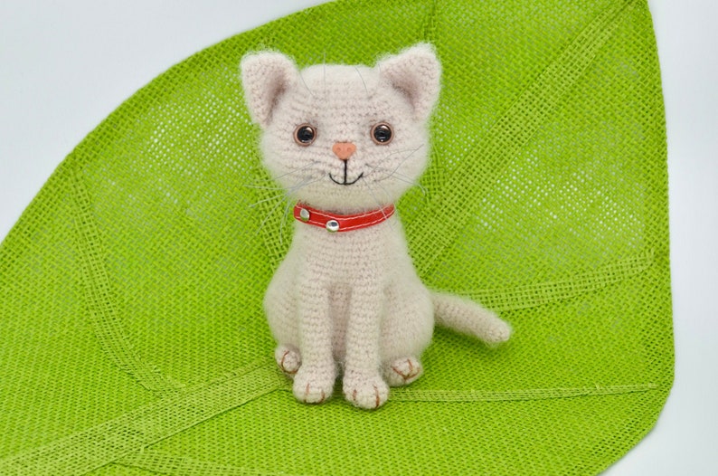 May include: A crocheted cat toy with a red collar. The cat is sitting on a green surface.