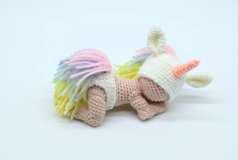 Puede incluir: Una mu&ntilde;eca de beb&eacute; unicornio de crochet con una melena y cola arco&iacute;ris. La mu&ntilde;eca lleva un gorro blanco de crochet con orejas y un cuerno.