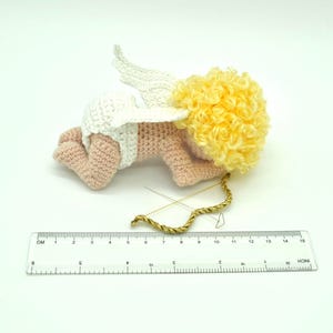 May include: A crocheted amigurumi cupid doll with pink skin, white wings, and a yellow curly wig. The doll is lying on its stomach with its head turned to the side. A gold bow and arrow is lying next to the doll.
