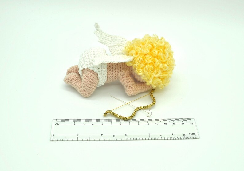 May include: A crocheted amigurumi cupid doll with pink skin, white wings, and a yellow curly wig. The doll is lying on its stomach with its head turned to the side. A gold bow and arrow is lying next to the doll.