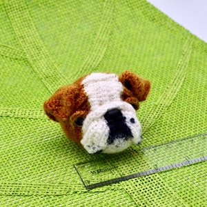 May include: A crocheted bulldog head with brown, white, and black fur. The bulldog is sitting on a green surface.
