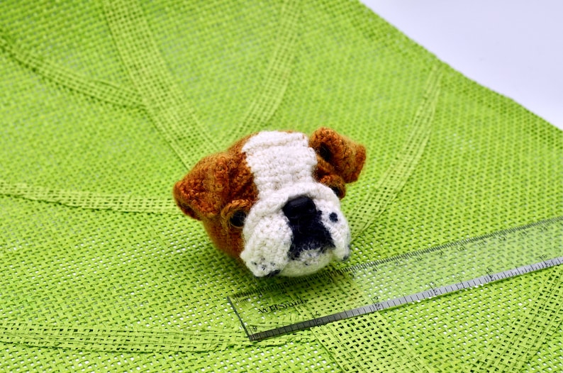 May include: A crocheted bulldog head with brown, white, and black fur. The bulldog is sitting on a green surface.