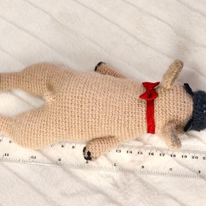 May include: A crocheted pug dog toy with black eyes and a red bow tie. The toy is lying on a white fluffy surface with a ruler measuring 25 inches.
