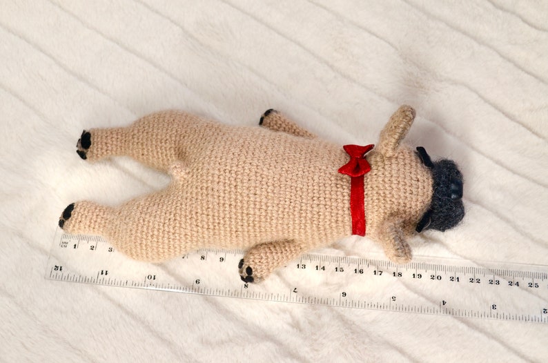 May include: A crocheted pug dog toy with black eyes and a red bow tie. The toy is lying on a white fluffy surface with a ruler measuring 25 inches.