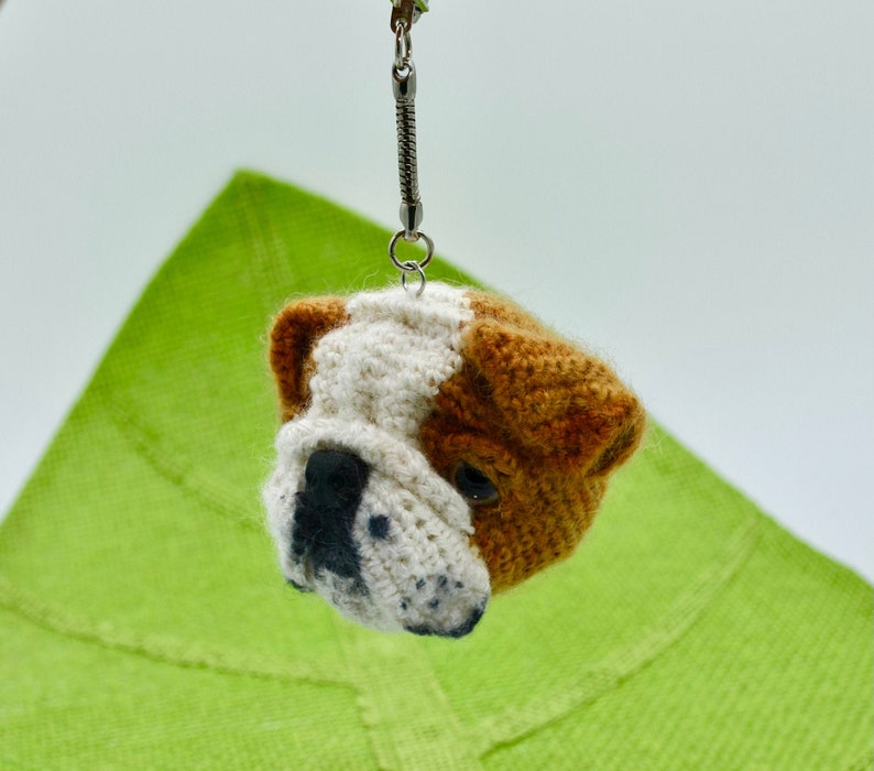 May include: A crocheted bulldog keychain with brown, white, and black fur. The keychain is attached to a silver chain.