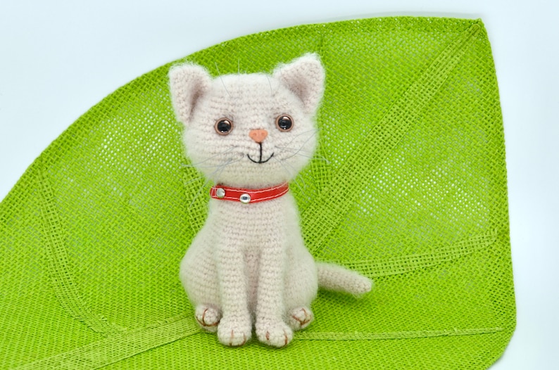 May include: A crocheted white cat with a red collar sitting on a green woven surface. The cat has brown eyes and a pink nose.