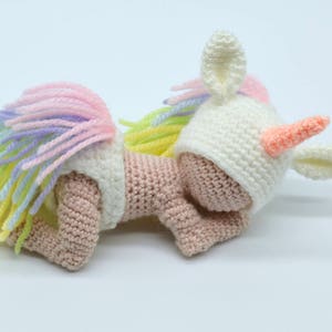 Puede incluir: Una mu&ntilde;eca de unicornio de crochet que lleva un gorro blanco con un cuerno y orejas rosas. La mu&ntilde;eca tiene un cuerpo rosa y una cola de arco&iacute;ris.
