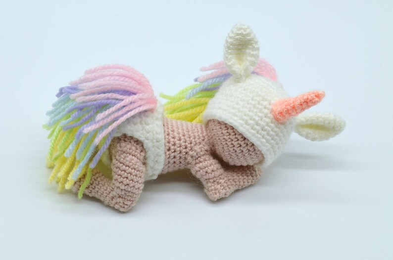 Puede incluir: Una mu&ntilde;eca de unicornio de crochet que lleva un gorro blanco con un cuerno y orejas rosas. La mu&ntilde;eca tiene un cuerpo rosa y una cola de arco&iacute;ris.