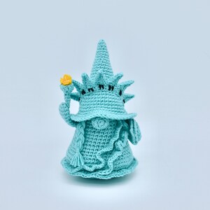 Puede incluir: Un gnomo de crochet con forma de la Estatua de la Libertad. El gnomo lleva un sombrero azul con una corona y sostiene una antorcha amarilla en su mano derecha.