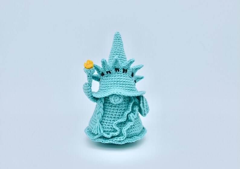 Puede incluir: Un gnomo de crochet con forma de la Estatua de la Libertad. El gnomo lleva un sombrero azul con una corona y sostiene una antorcha amarilla en su mano derecha.