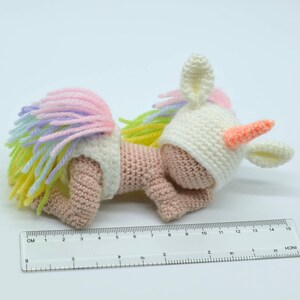 Puede incluir: Un peque&ntilde;o juguete de unicornio de crochet con una melena y cola arco&iacute;ris. El unicornio lleva un gorro blanco de crochet con un cuerno rosa y est&aacute; tumbado de lado.