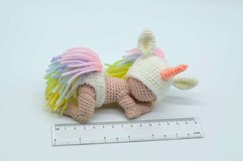 Puede incluir: Un peque&ntilde;o juguete de unicornio de crochet con una melena y cola arco&iacute;ris. El unicornio lleva un gorro blanco de crochet con un cuerno rosa y est&aacute; tumbado de lado.