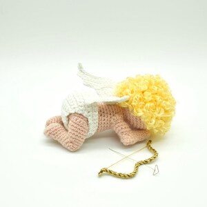 May include: A crocheted amigurumi cupid doll with a white diaper, pink body, white wings, and yellow curly hair. The cupid is lying on its stomach with its bow and arrow beside it.