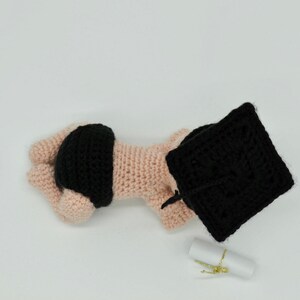 May include: A crocheted baby doll wearing a black graduation cap and a black diaper. The doll is lying on its side with its head resting on a white surface. A small white scroll is in the background.