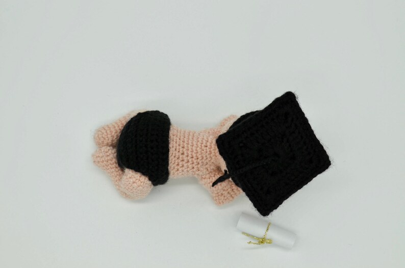 May include: A crocheted baby doll wearing a black graduation cap and a black diaper. The doll is lying on its side with its head resting on a white surface. A small white scroll is in the background.