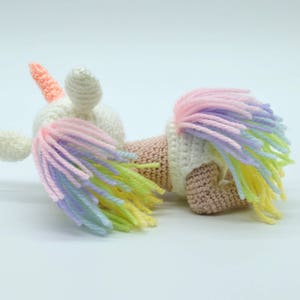 Puede incluir: Un juguete de unicornio de crochet blanco con un cuerno rosa y una crin arco&iacute;ris. El unicornio est&aacute; acostado de lado con la cabeza girada.