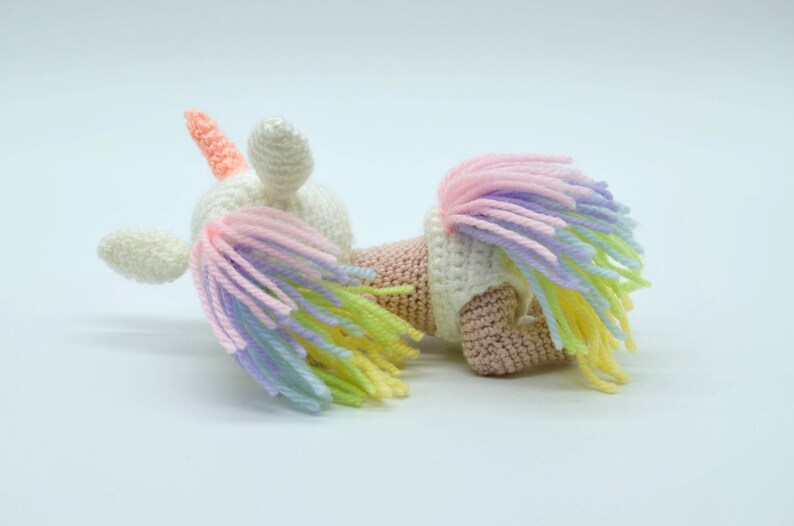 Puede incluir: Un juguete de unicornio de crochet blanco con un cuerno rosa y una crin arco&iacute;ris. El unicornio est&aacute; acostado de lado con la cabeza girada.