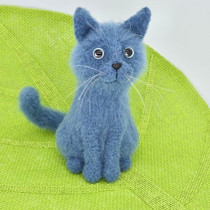 May include: A blue knitted cat toy with white whiskers and large black eyes. The cat is sitting on a green woven mat.