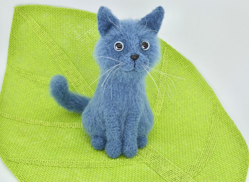 May include: A blue knitted cat toy with white whiskers and large black eyes. The cat is sitting on a green woven mat.