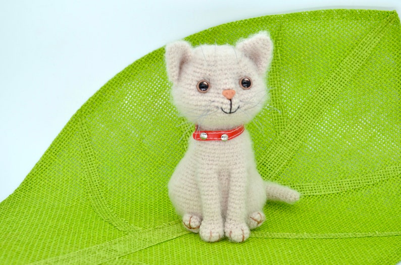 May include: A crocheted white cat with a red collar sits on a green crocheted surface. The cat has black eyes and a pink nose.
