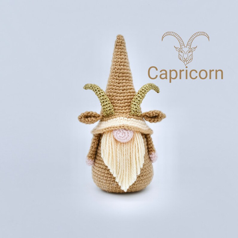 May include: Crocheted Capricorn gnome with a brown hat, gold horns, and a cream-coloured beard. The gnome is standing on a white background. The text "Capricorn" is written in brown above the gnome.