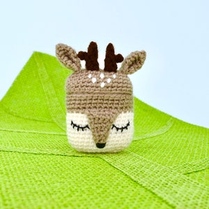 May include: A crocheted deer-shaped container with closed eyes, brown antlers, and white spots. The container is beige and white, set against a bright green textured background. The deer has a black nose and closed eyes.