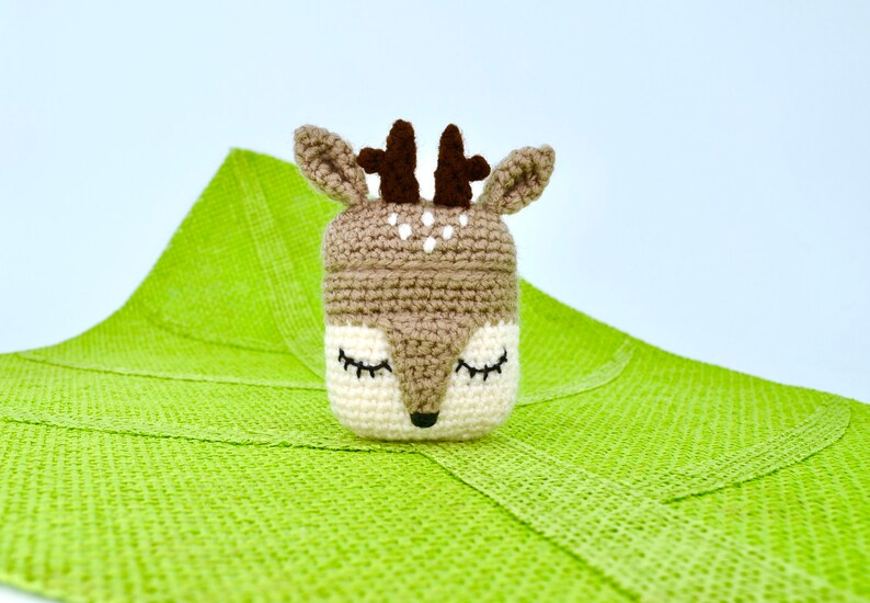 May include: A crocheted deer-shaped container with closed eyes, brown antlers, and white spots. The container is beige and white, set against a bright green textured background. The deer has a black nose and closed eyes.