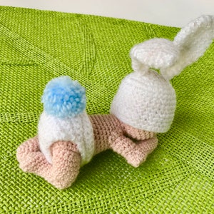 Puede incluir: Un juguete de conejo de crochet con un cuerpo rosa claro, un gorro y orejas blancas y un pompón azul claro en la parte superior del gorro. El conejo está acostado sobre una superficie tejida verde.