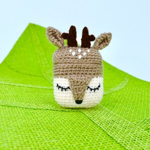 May include: A handmade crochet deer toy with closed eyes, brown antlers, and a white and brown face. The deer is set against a bright green textured background. The toy is likely a decorative item or a child's toy.