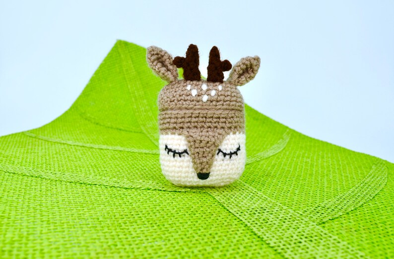 May include: A handmade crochet deer toy with closed eyes, brown antlers, and a white and brown face. The deer is set against a bright green textured background. The toy is likely a decorative item or a child's toy.