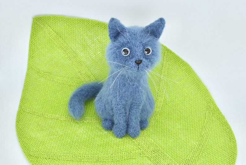 May include: A blue knitted cat toy with white whiskers sitting on a green leaf-shaped mat.