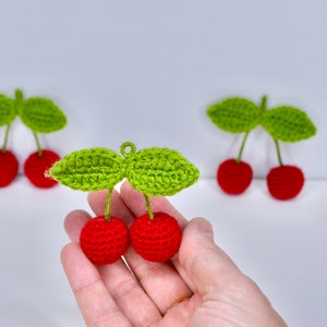 Könnte beinhalten: Drei gehäkelte Kirschen mit grünen Blättern. Die Kirschen sind rot und die Blätter sind grün. Die Kirschen werden in einer Hand gehalten.