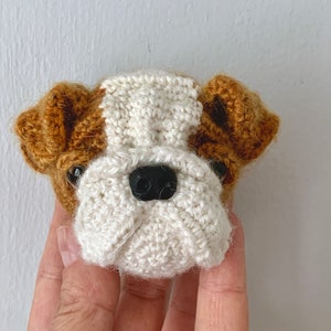 May include: A crocheted bulldog head with brown ears and a white face. The bulldog has black eyes and a black nose.