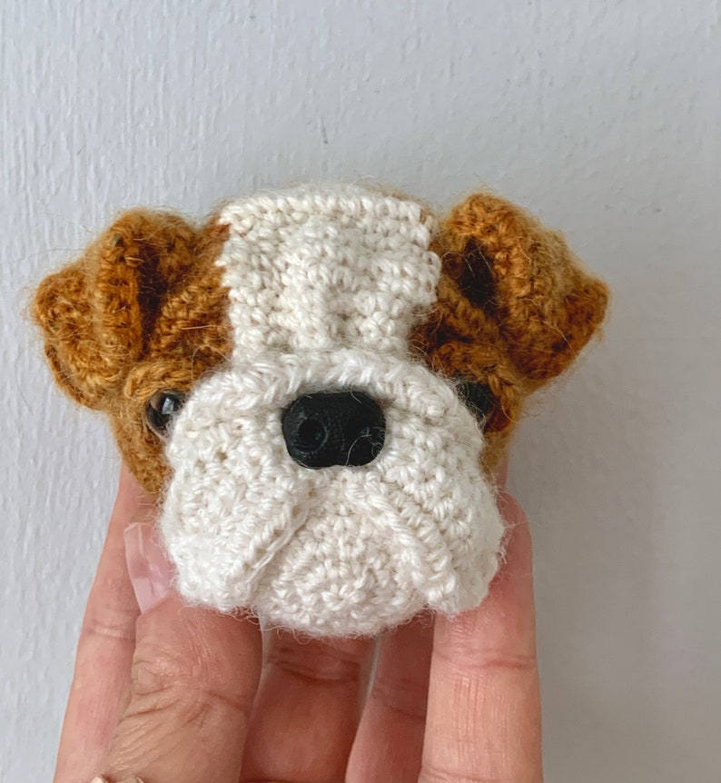 May include: A crocheted bulldog head with brown ears and a white face. The bulldog has black eyes and a black nose.