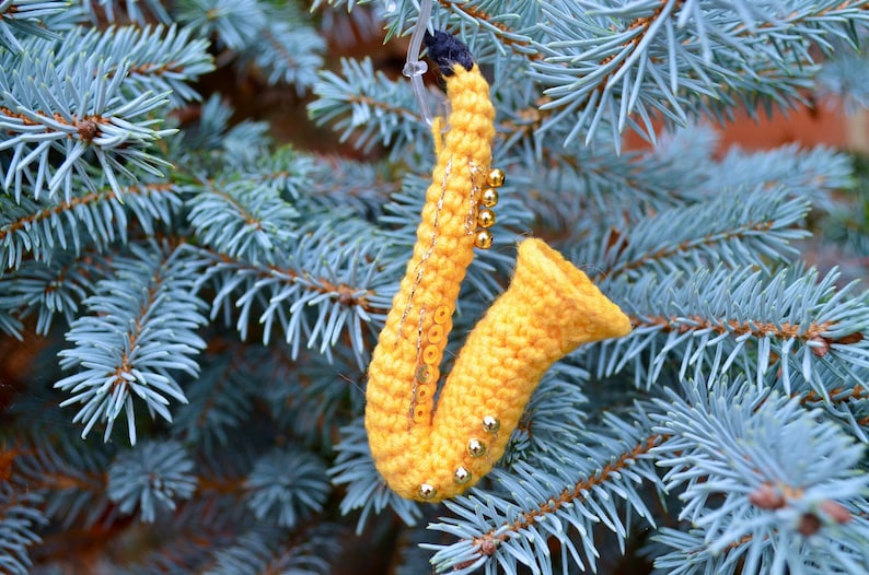 May include: A yellow crocheted saxophone ornament with gold beads hanging from a blue spruce tree.