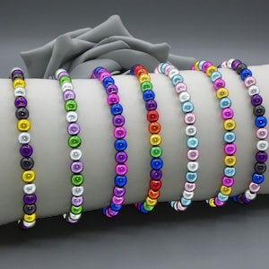 May include: Seven beaded bracelets arranged in a row, each featuring a unique sequence of glossy, spherical beads in vibrant colours including yellow, purple, green, pink, blue, orange, and white, all strung together on a flexible cord.