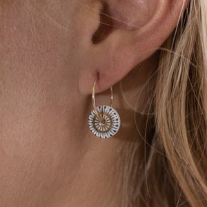 May include: Gold hoop earrings with a white, blue, and gold circular pendant. The pendant features a detailed geometric design. The earrings are shown on a person's ear, highlighting the jewelry's design and style.