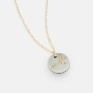 Könnte beinhalten: Halskette mit goldfarbener Kette und einem runden, hellgrünen Anhänger. Der Anhänger hat ein goldfarbenes, abstraktes Design. Die Halskette ist ein schlichtes, elegantes Schmuckstück.